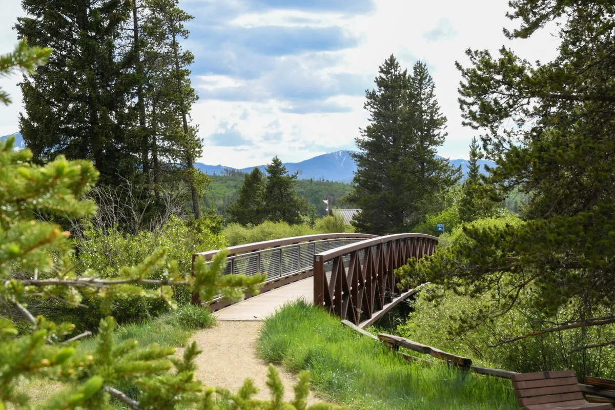 A peaceful exterior view featuring a charming wooden bridge surrounded by lush trees and a scenic mountain backdrop. The area is ideal for walking, biking, or enjoying nature, suggesting easy access to trails and family-friendly outdoor adventure.