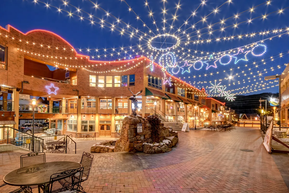 An inviting downtown scene with twinkling string lights, decorative stars, a stone fountain, and an outdoor seating area. People can be seen strolling, socializing, and enjoying the lively ambiance, suggesting nightlife and the festive resort-town atmosphere.