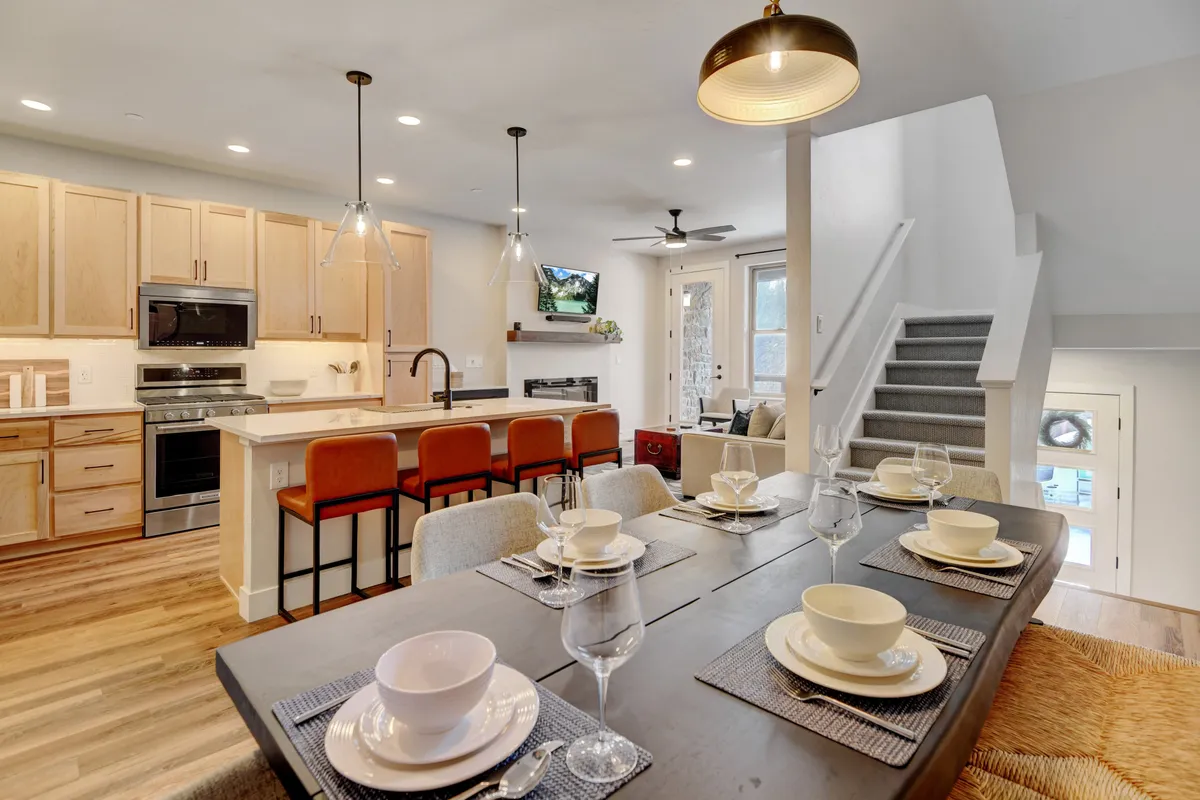 The image showcases a spacious, sunlit open-concept living area featuring a beautifully set dining table, a modern kitchen island with bar stools, and a cozy living space with an inviting sofa—all styled for comfort and connection. The atmosphere is warm, with hints of mountain-inspired decor that reflect the Colorado setting.