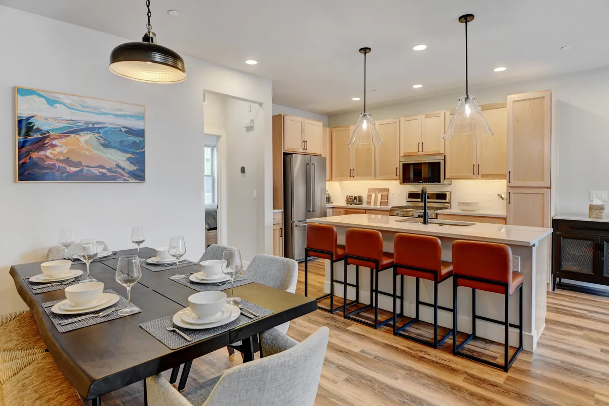 A dining table beautifully set for a shared meal stands adjacent to a kitchen island with bar stools, highlighted by colorful artwork on the wall. The space radiates warmth and hospitality, ideal for family feasts or casual gatherings, and visually links the kitchen's function with the joy of shared dining experiences.