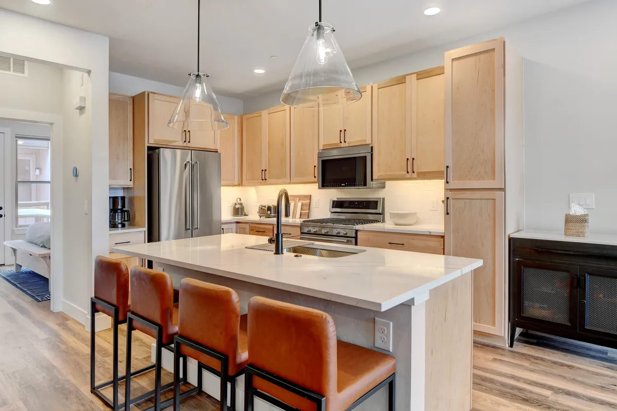 This image showcases the heart of the kitchen space, featuring a spacious island counter with stylish bar stools, modern kitchen appliances, and warm wood cabinetry. The setup invites cooking, gathering, and entertaining, highlighting both the premium features and the social atmosphere of the culinary space.