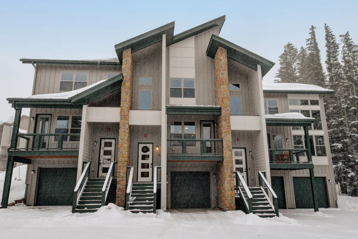 This exterior image highlights the property's stone pillars, balconies, and large windows, all set against a snowy mountain landscape. The steps leading up to the entrance are dusted with snow, and the scene evokes a sense of adventure, welcoming guests into a mountain getaway.