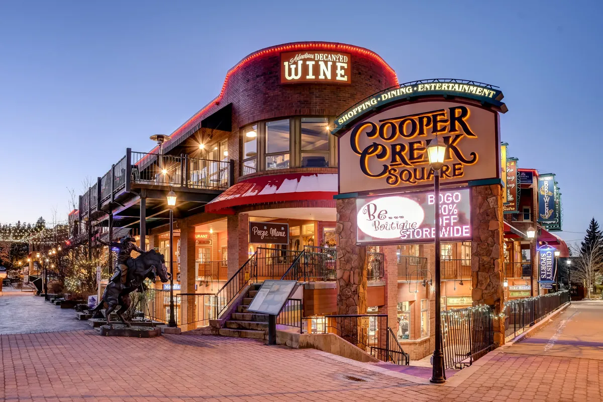 A vibrant downtown streetscape featuring the Cooper Creek Square sign, inviting outdoor seating areas, and storefronts for wine shops and restaurants. The scene is lively, suggesting year-round dining and entertainment in downtown Winter Park.
