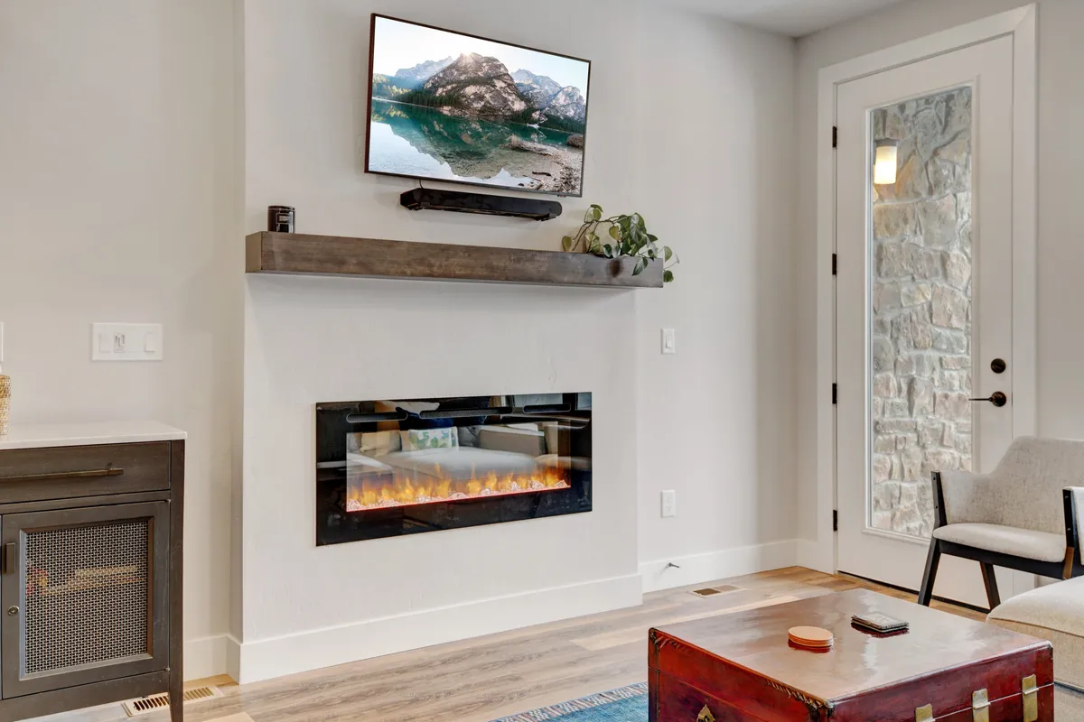 A modern, inviting living area featuring a striking electric fireplace, a wall-mounted television displaying mountain scenery, and a stylish decorative shelf with plants. Plush furnishings and a cozy rug set the scene for relaxation, socializing, and entertainment.