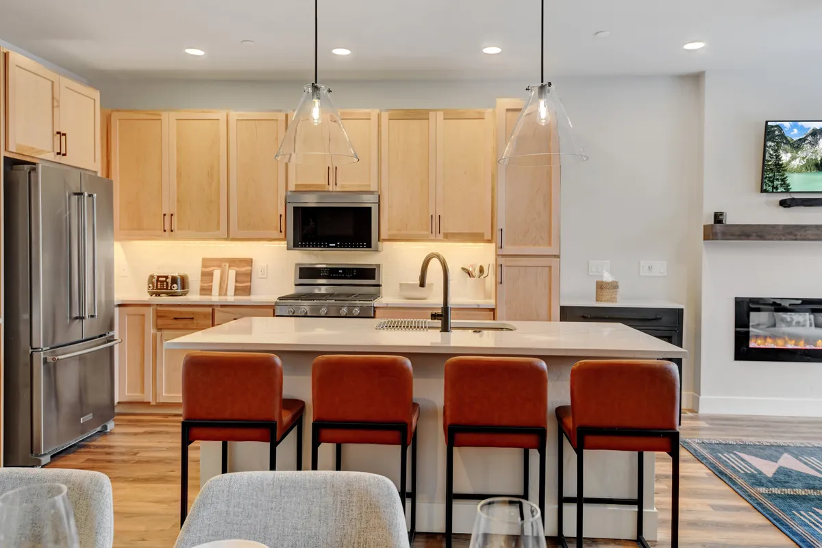 A welcoming open-plan kitchen extends into a cozy living area, with an island countertop, bar stools, high-end appliances, pendant lighting, and a fireplace in the background. The visual flow connects culinary prep with socializing and relaxation, embodying the seamless transition between cooking, dining, and lounging.